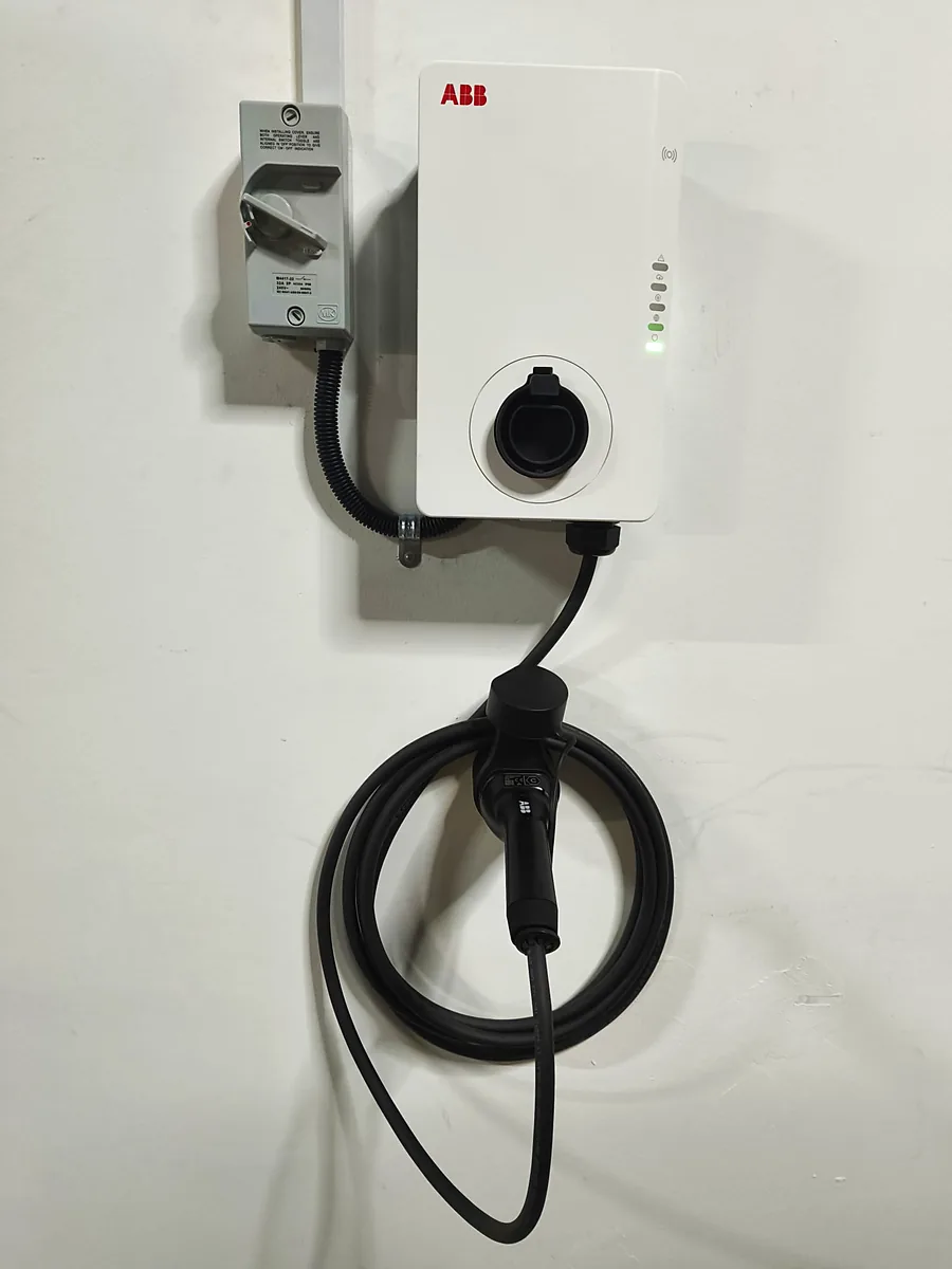 A white ABB charger beside its switch enclosure, the cable stowed on a holster below.