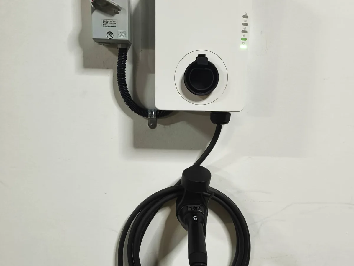 A white wall-mounted AC charger with status LEDs, wired in conduit with its Type 2 cable coiled below