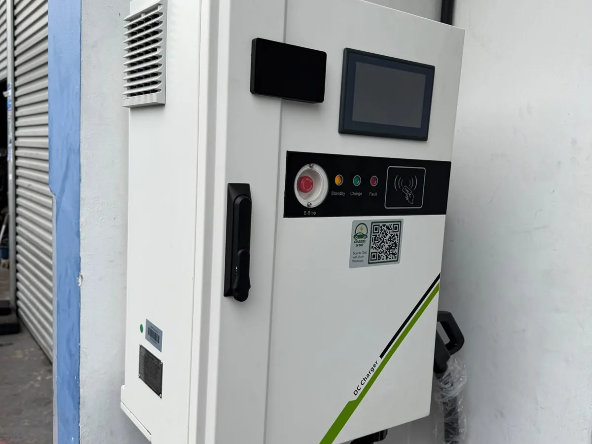 A white DC fast-charging cabinet with a touch display, a red emergency-stop button, a row of status indicators and a contactless reader, standing in front of a roller shutter.