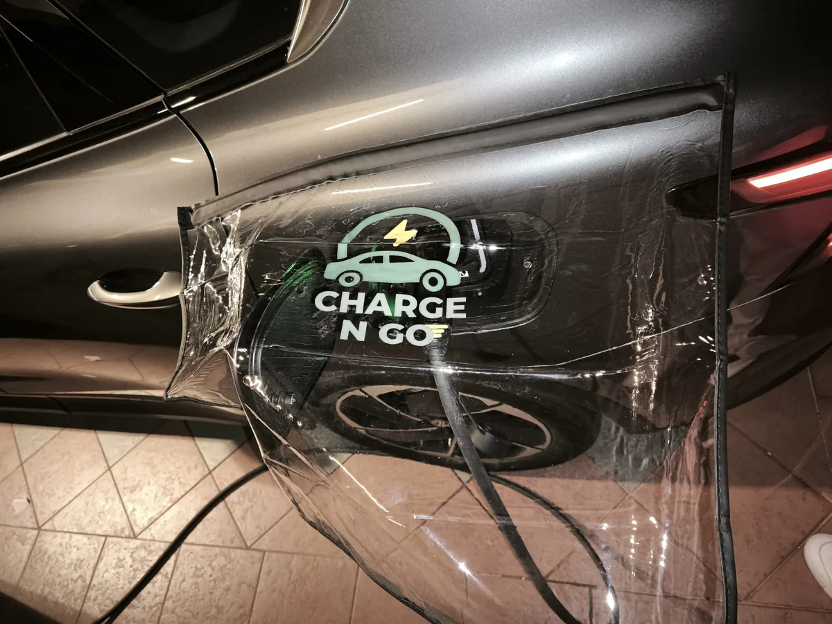 A clear Charge N Go rain shield clipped over the charging port of a dark car, tiled floor below.