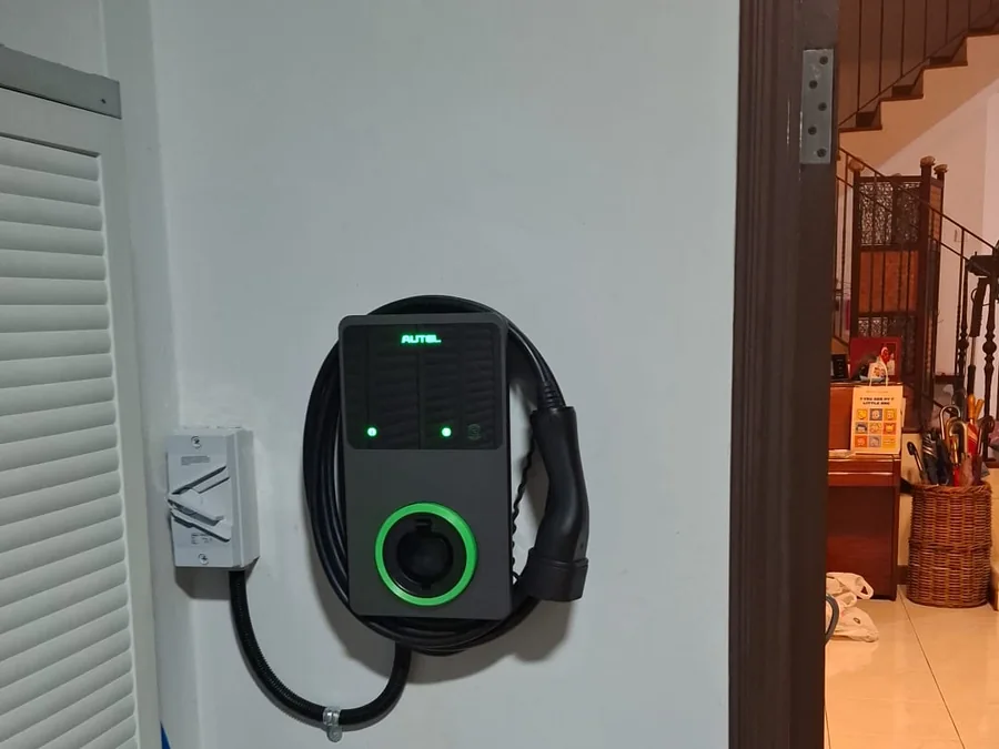 An Autel wall charger with a green ring mounted on a corridor wall, its cable coiled beneath and a doorway visible beyond.