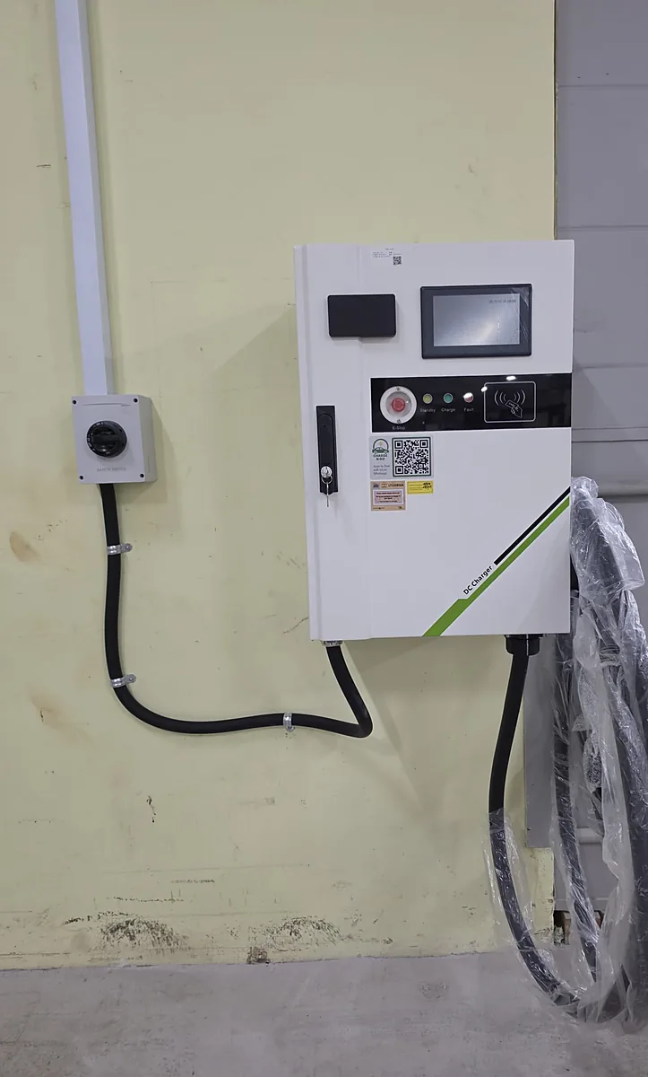 A wall-mounted DC EV charger with a display screen still under protective film, an emergency-stop button and status lights, its tethered cable coiled in protective wrap, fed by black conduit from a wall-mounted safety switch.