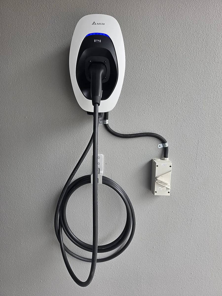 A white wall-mounted EV charger with a blue status ring and its connector docked, the cable looped on a wall hook and the supply running in flexible conduit to a labelled isolator switch alongside.