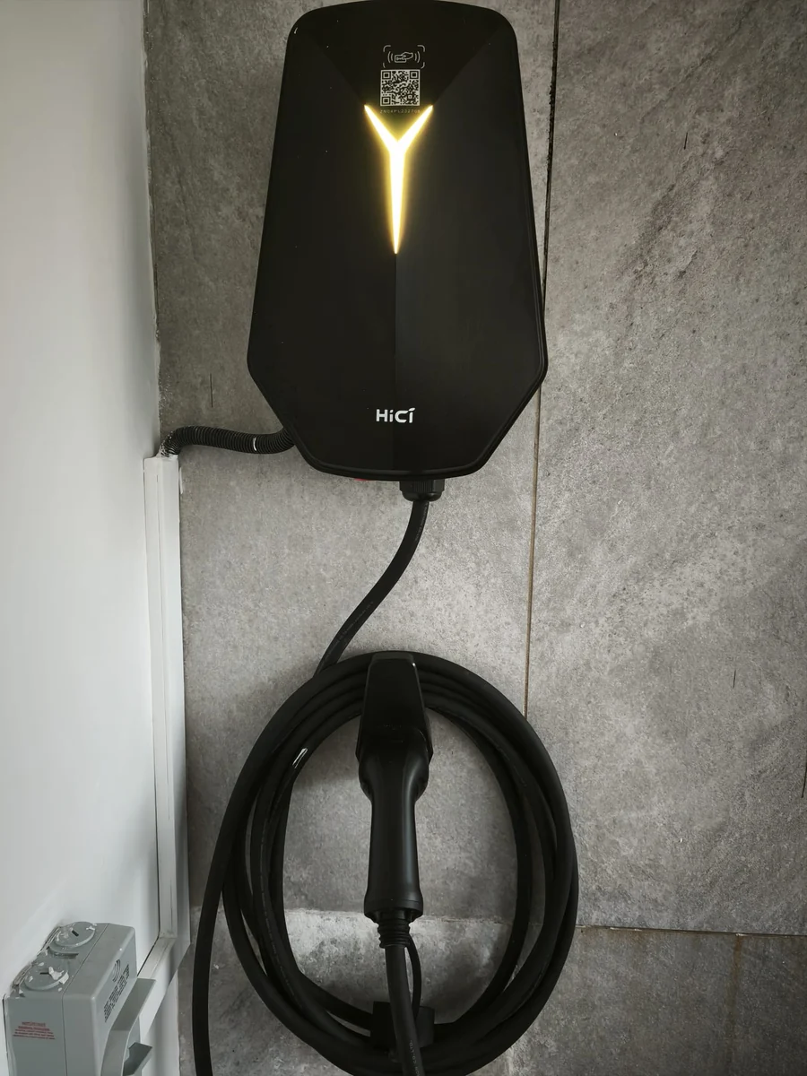A black faceted HiCi charger with its Y-shaped light lit amber, on a grey stone-tiled wall with the cable coiled on a holster below.