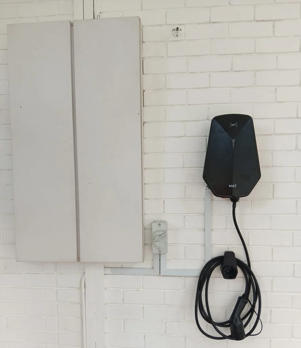 A black faceted HiCi charger on a white painted brick wall beside a tall white cabinet, an isolator below it and the connector stowed on a holster.