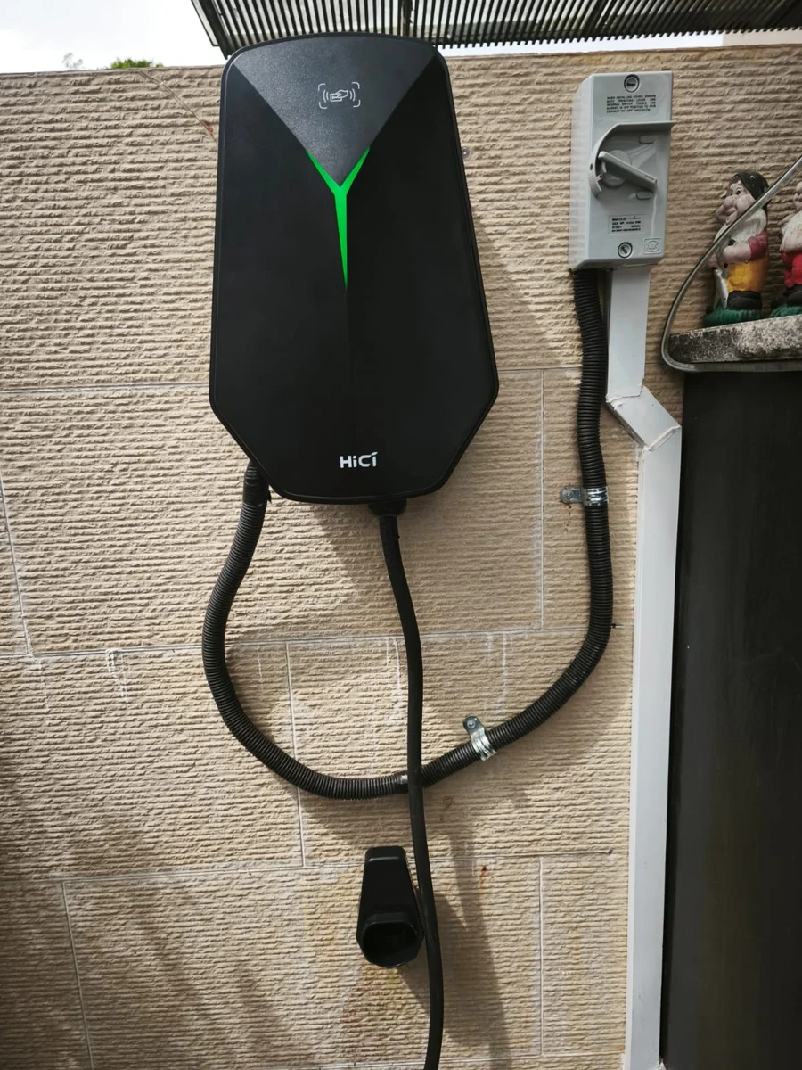 A black faceted HiCi charger with its Y light lit green, mounted on a textured wall under a slatted screen with an isolator beside it.