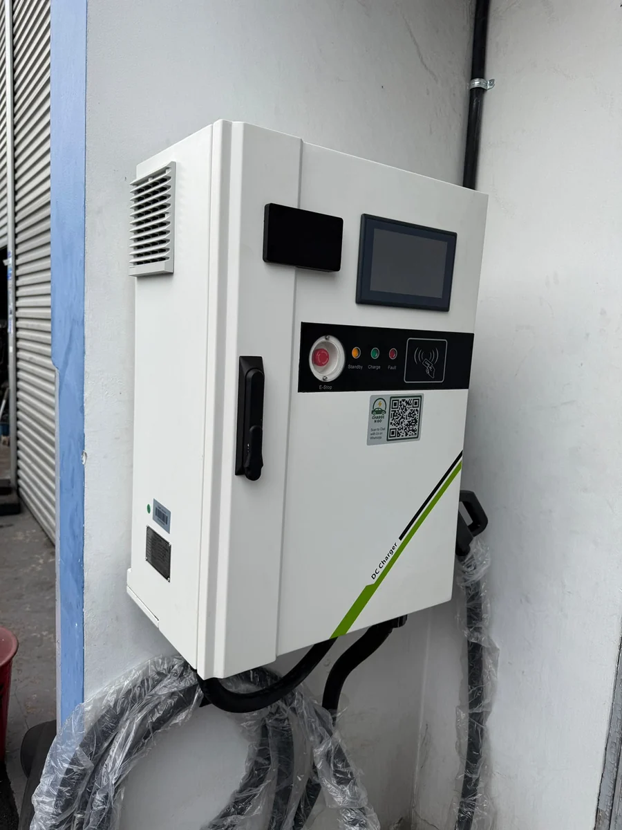A white wall-mounted DC fast charger with a vented side, touchscreen and QR label, mounted beside a roller shutter.