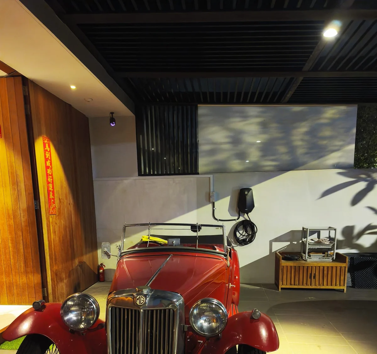 A covered garage at night with a red vintage MG sports car parked beside a wall-mounted charger, timber panelling and a fire extinguisher behind.