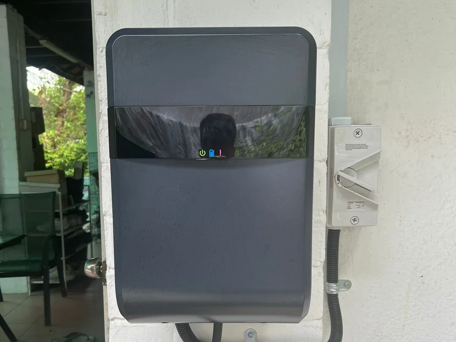 A compact black wall charger with a reflective front panel and status indicators, mounted on a white wall with conduit entering from below.