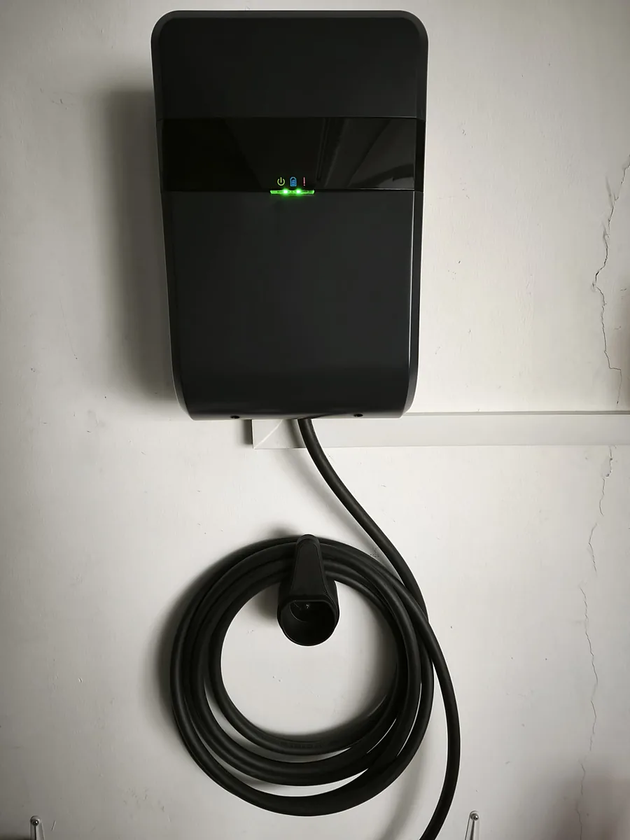 A slim black wall charger with a green LED status bar, mounted on a plain white interior wall.