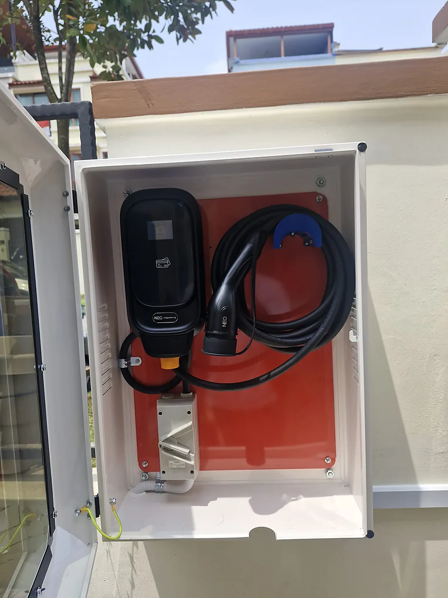 A black EV charger on a red backboard inside a white outdoor enclosure, its coiled lead and connector resting on a blue hook, with a white weatherproof switch enclosure and conduit below.