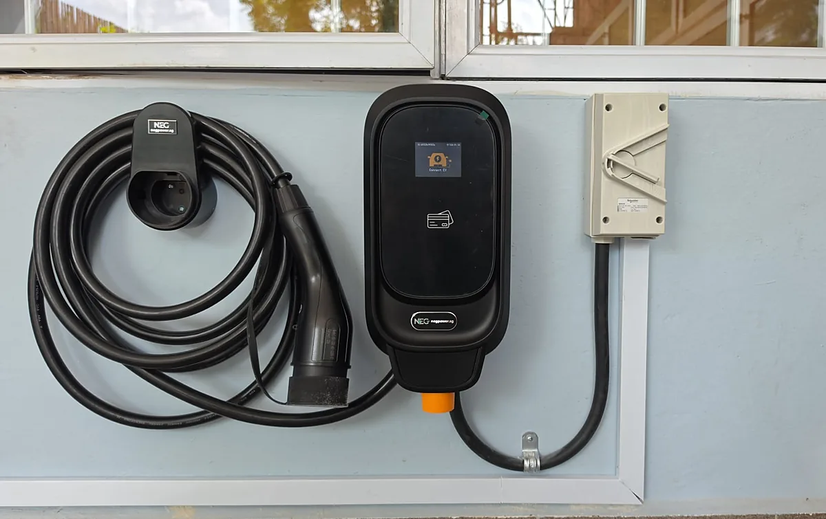 A black wall-mounted EV charger with a lit display reading 'Connect EV', its coiled lead hung on a holster to the left and a cream enclosed isolator to the right, on a pale blue painted wall below a run of windows.