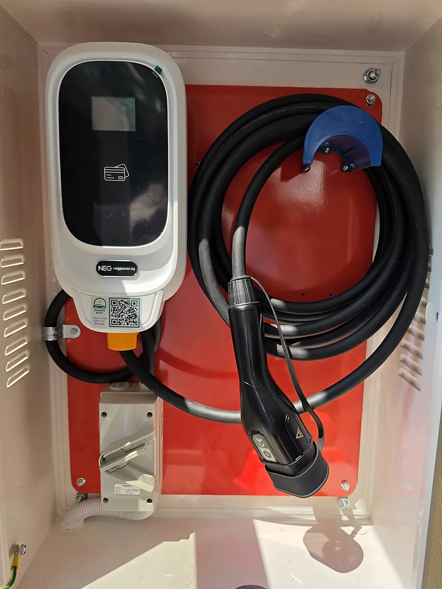 A white NEG wall-mounted EV charger on a red backboard inside a white louvred cabinet, its coiled lead over a blue hook and the Type 2 connector docked in a holster below.