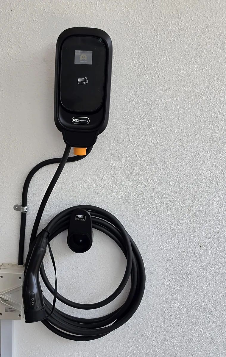 A black wall-mounted NEG EV charger on a white textured wall, its coiled charging lead hung on a holster below and the supply cable running down to a white enclosure at the foot of the wall.