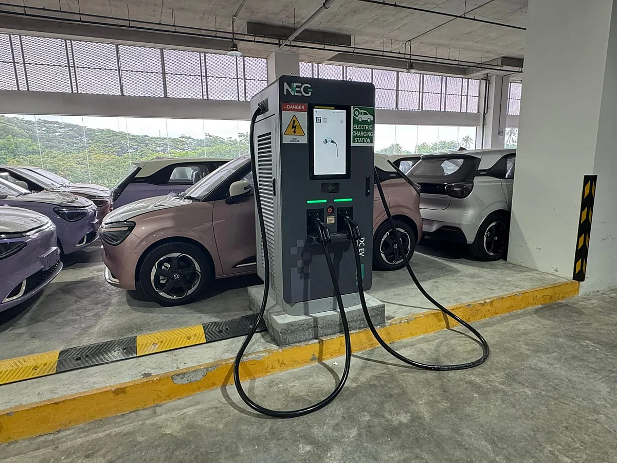 A free-standing DC charging cabinet on a concrete plinth in a covered car park, a red high-voltage label and a green "Electric Charging Station" sign either side of its touchscreen, both cables looped across the floor.