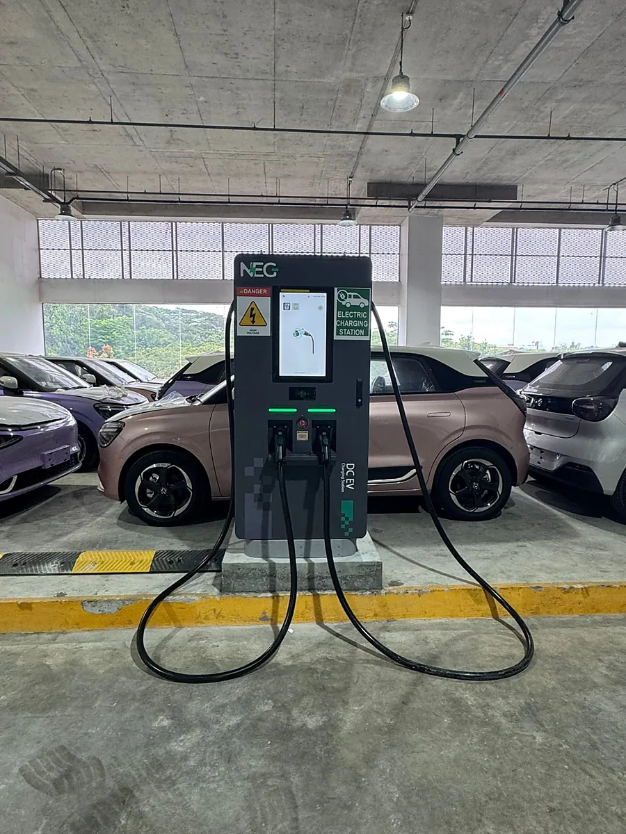 A floor-standing DC charger with two tethered cables and a lit colour screen, its green status bars showing above the connector holsters, mounted on a concrete plinth in a covered car park.
