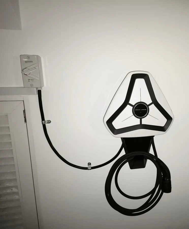 A white triangular Omoda charger on an interior wall beside a louvred door, an isolator to the left and the cable coiled beneath.