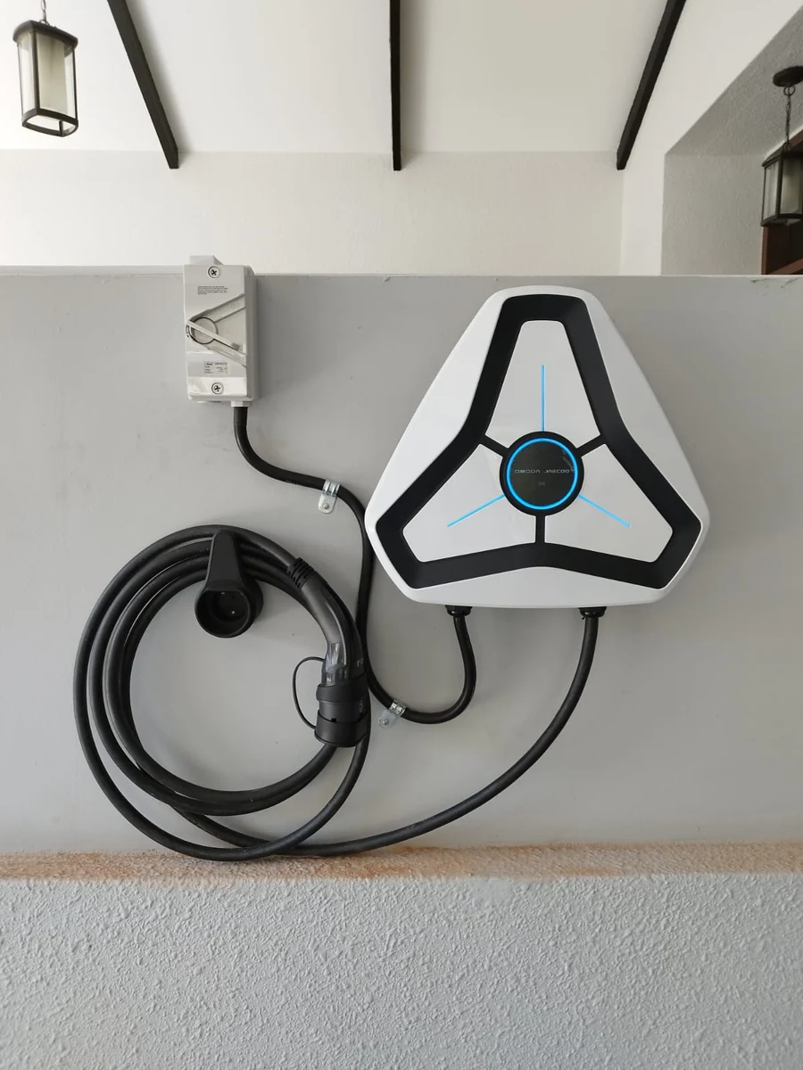 A white triangular Omoda charger on a rendered wall beneath a white beamed ceiling, an isolator above left and the tethered cable coiled on its holster.