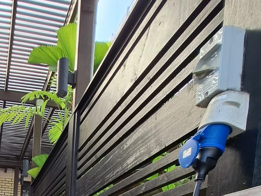 A weatherproof industrial socket outlet fixed to a timber screen wall under a carport, with a blue commando plug inserted and the cable dropping away below.