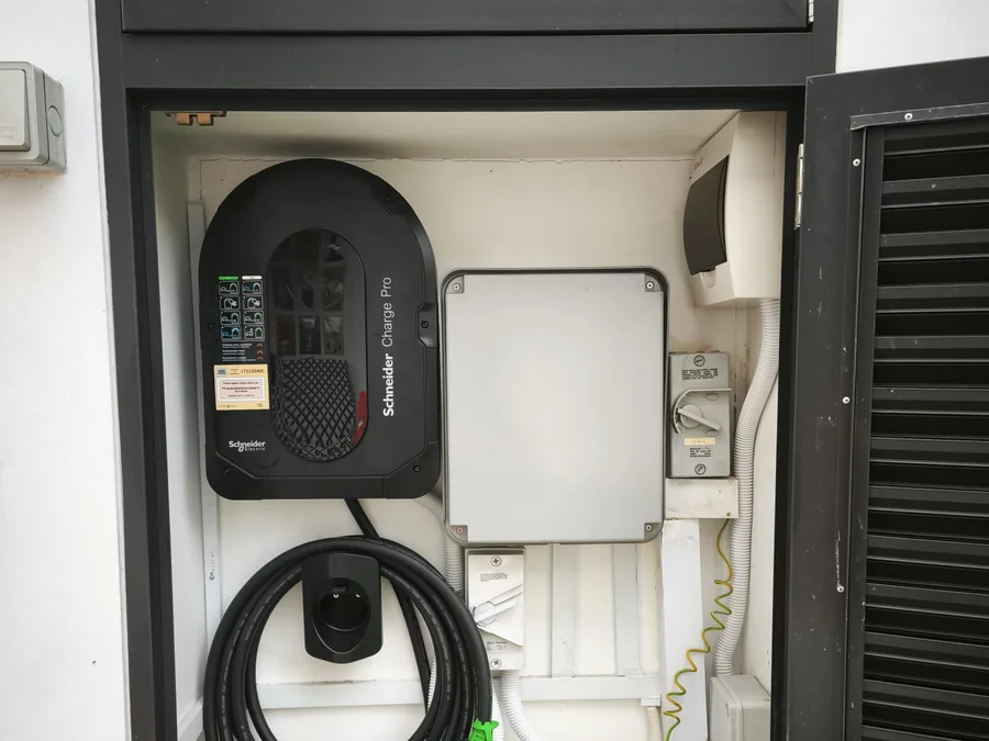 A Schneider Charge Pro wall charger mounted inside a recessed wall cabinet beside a louvred vent, its cable coiled on a holster below.
