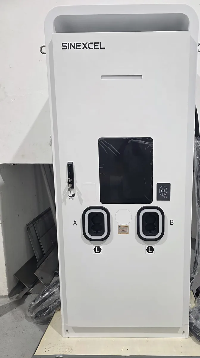 A white Sinexcel floor-standing EV charging cabinet photographed straight on indoors, its display still covered in protective film and both connector sockets closed and unused, with an installer working at an adjacent panel to the right.