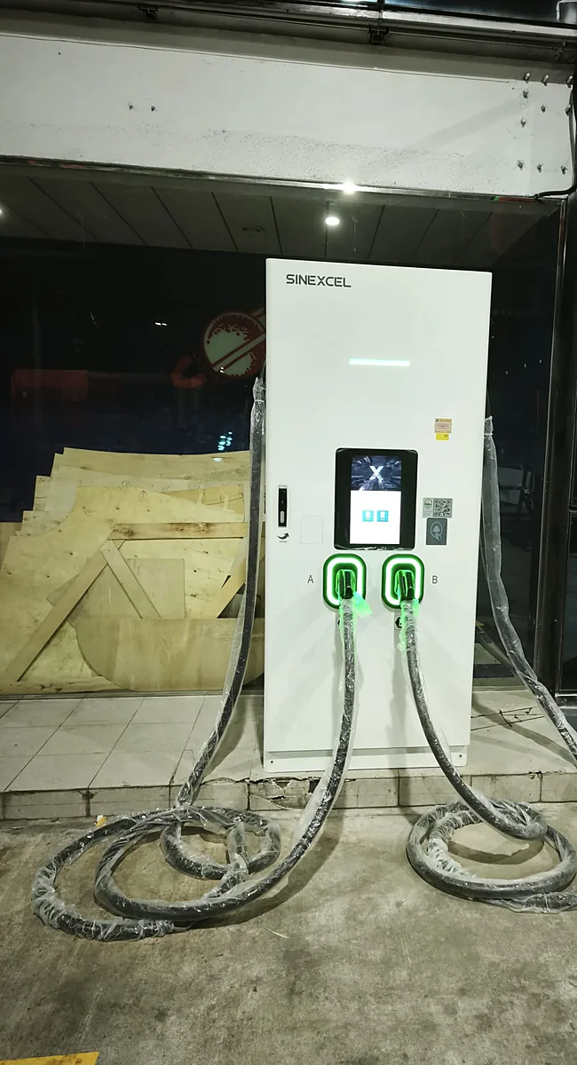 A floor-standing white DC charging cabinet with a lit colour screen and two green-lit connector holsters, its cables still in factory wrapping and coiled on the concrete beside it.